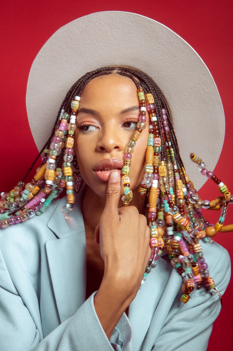 RiRi, editorial model, in hand-beaded braids and a cream wide-brim hat with a powder-blue blazer on a red backdrop