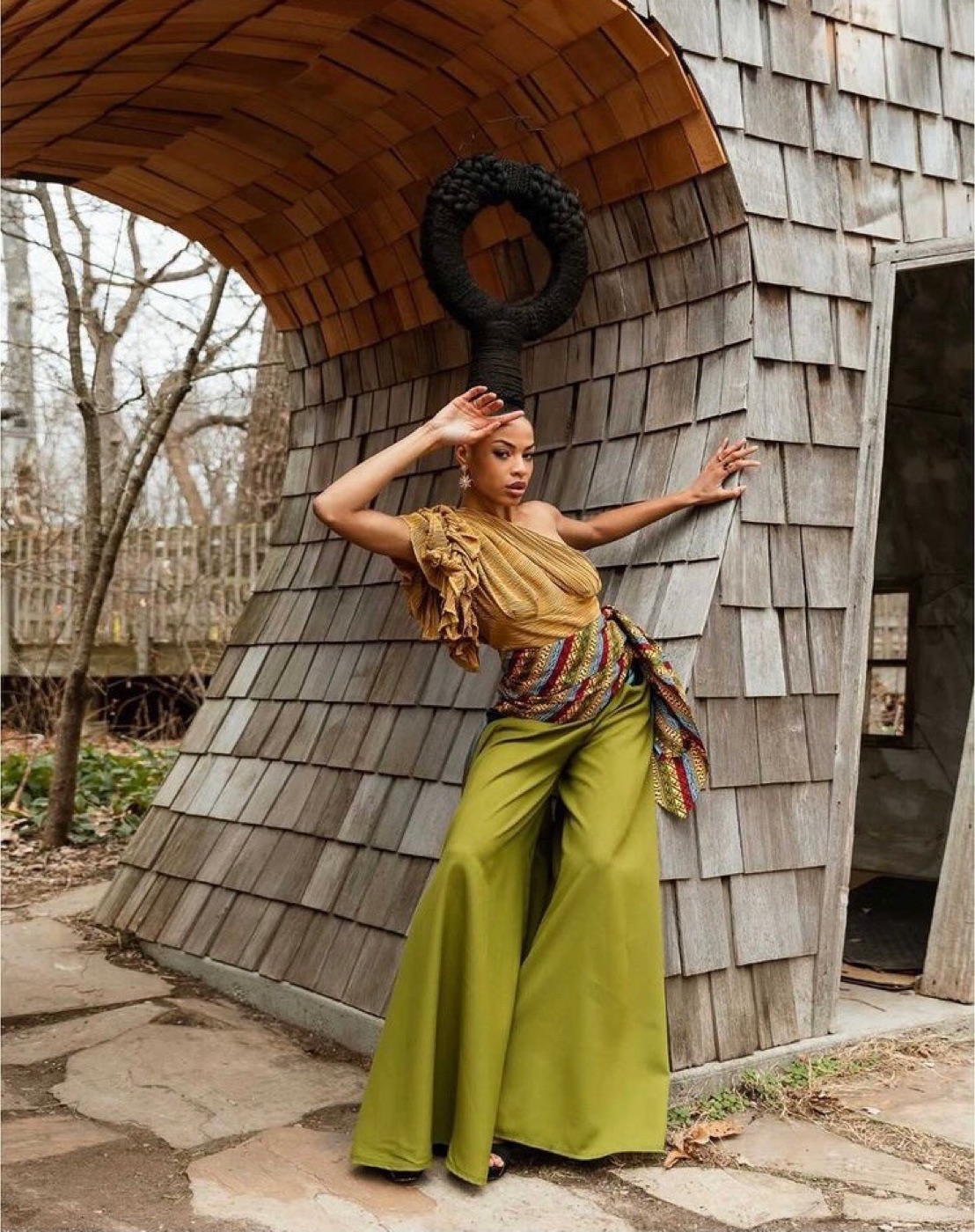RiRi, fashion model, in a mustard one-shoulder top and chartreuse wide-leg pants against a wood-shingle structure